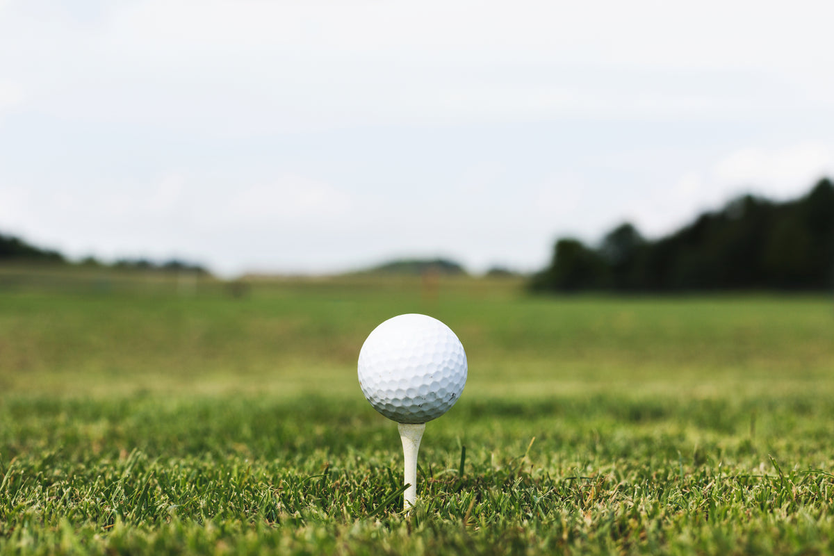 Im Vordergrund ist ein Golfball abschlagbereit auf einem Tee positioniert. Dahinter sieht man nichts als Wiese und ein paar Bäume. Gerade wenn beim Golf weit und breit kein Wasserspender in der Nähe ist, sind unsere 720°DGREE-Trinkflaschen einfach perfekt